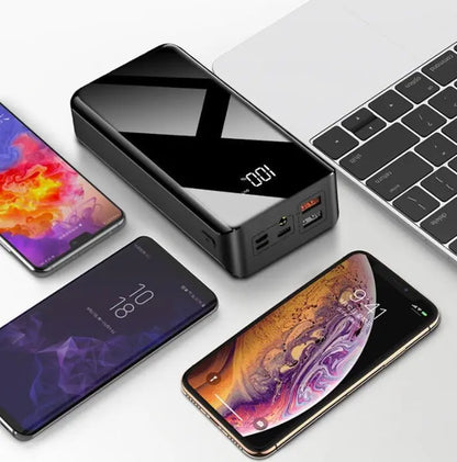Black 30000mAh power bank with digital display and multiple USB ports charging three smartphones next to a laptop keyboard