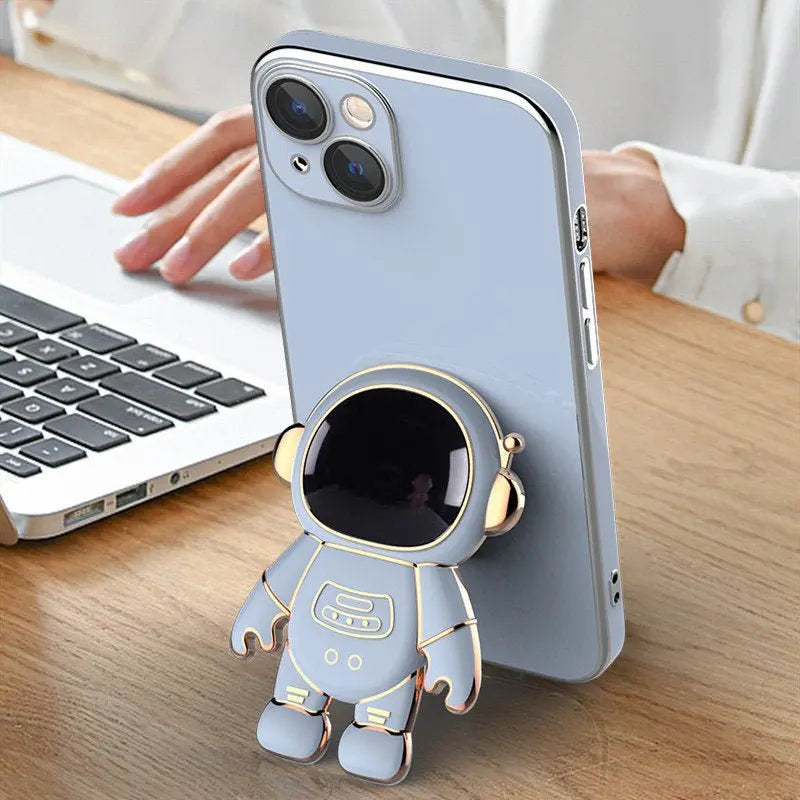 Light blue iPhone with 3D astronaut phone case featuring electroplating bracket standing on wooden desk next to laptop