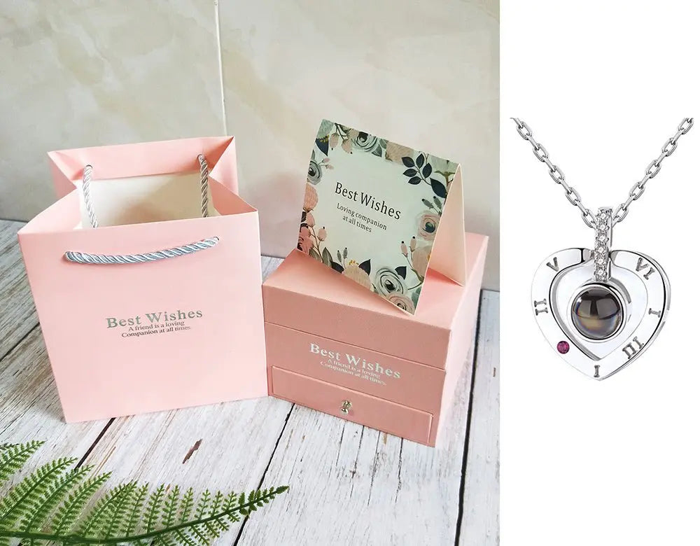 Elegant silver heart-shaped necklace with Roman numerals and a small red gem, displayed with pink gift box and card set