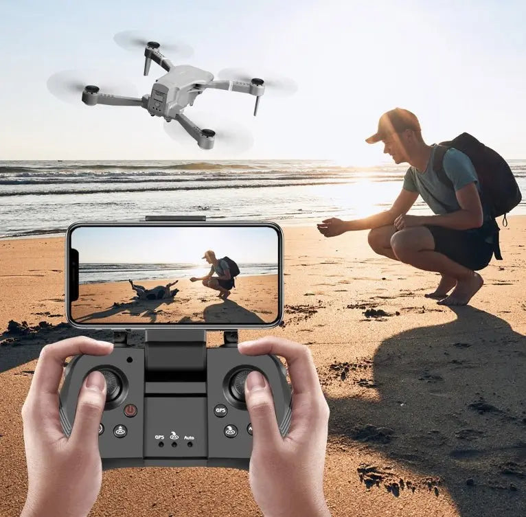 Hands holding remote controller with smartphone showing beach scene, folding drone flying over sandy shore at sunset