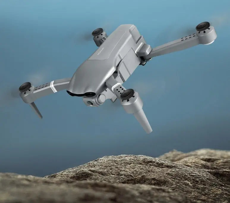 Grey folding drone in flight above rocky terrain with visible front camera and extended arms