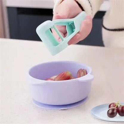 Hand holding mint green fruit slicer cutter above purple bowl with halved grapes inside on kitchen counter