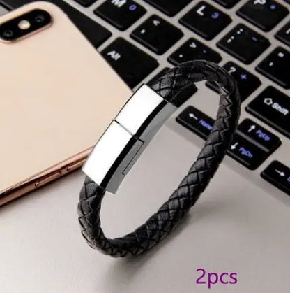 Black braided leather bracelet USB charging cable with silver magnetic clasp placed on laptop keyboard next to smartphone