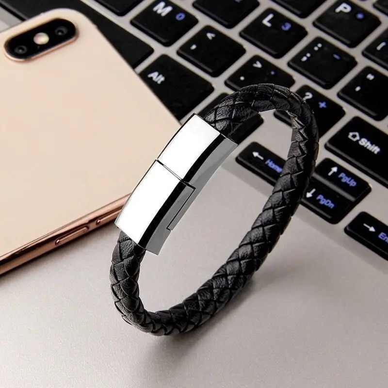 Black braided leather bracelet charger with silver magnetic clasp displayed on laptop keyboard next to smartphone