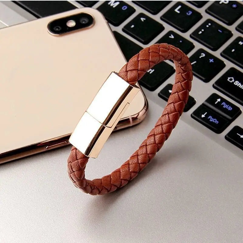 Brown braided leather bracelet USB charging cable with gold metallic magnetic clasp placed on a laptop keyboard next to a smartphone