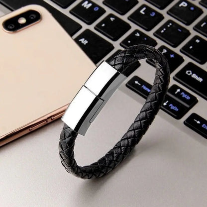 Black braided leather bracelet USB charging cable with silver magnetic clasp, placed on laptop keyboard beside smartphone