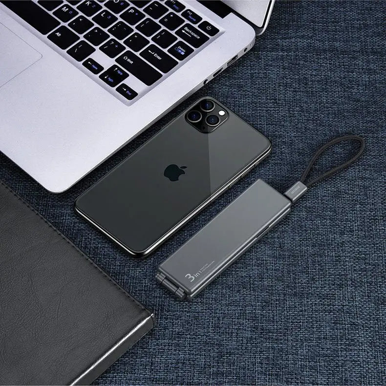 Portable Multifunctional One For Three Charger in dark grey with wrist strap placed beside a smartphone and laptop keyboard