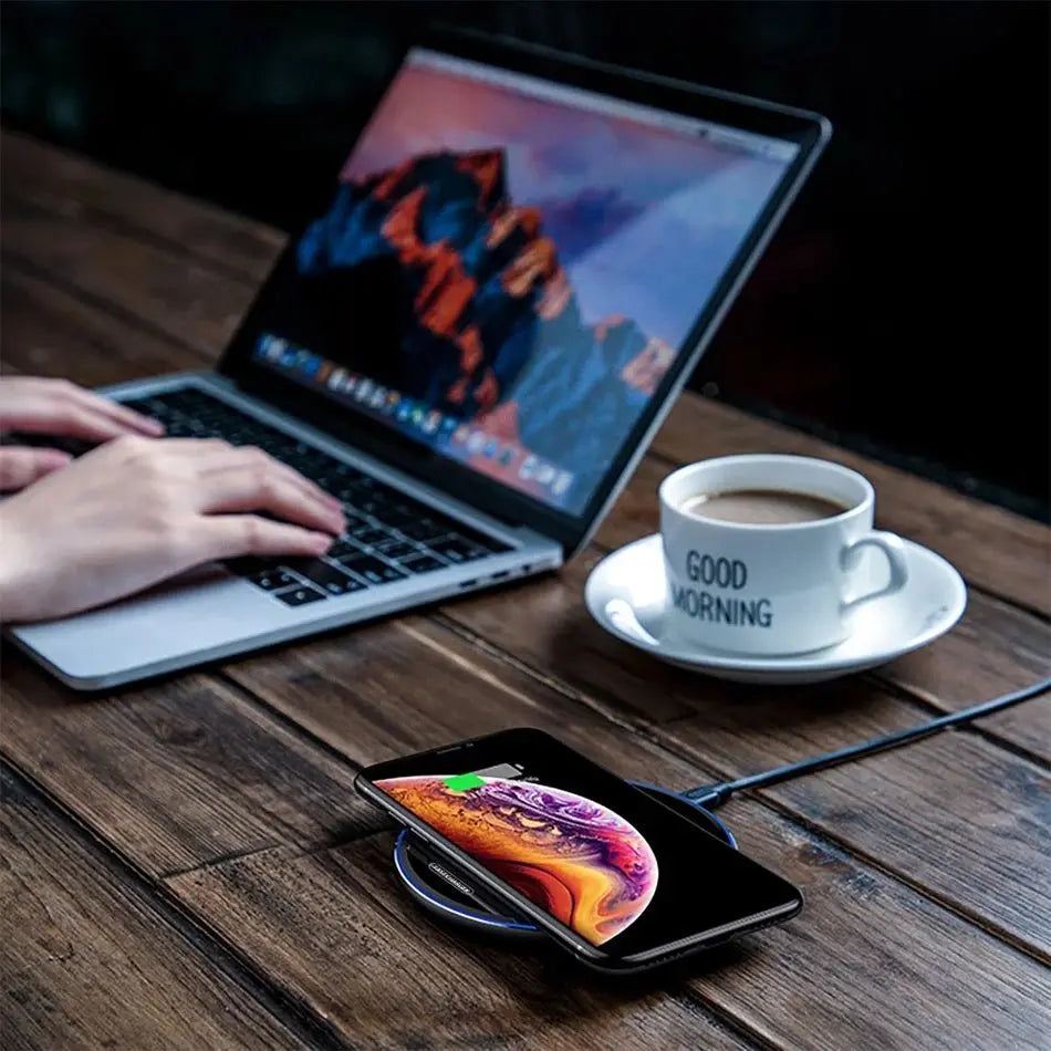 Wireless charger pad with smartphone charging, laptop in background, and coffee cup on wooden table surface